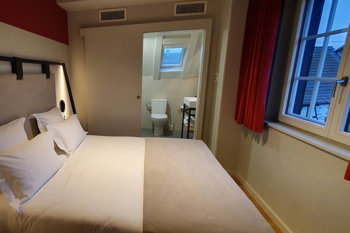 Una cama bien tendida con sábanas blancas, cabecera con estructura negra y una ventana con cortinas rojas.