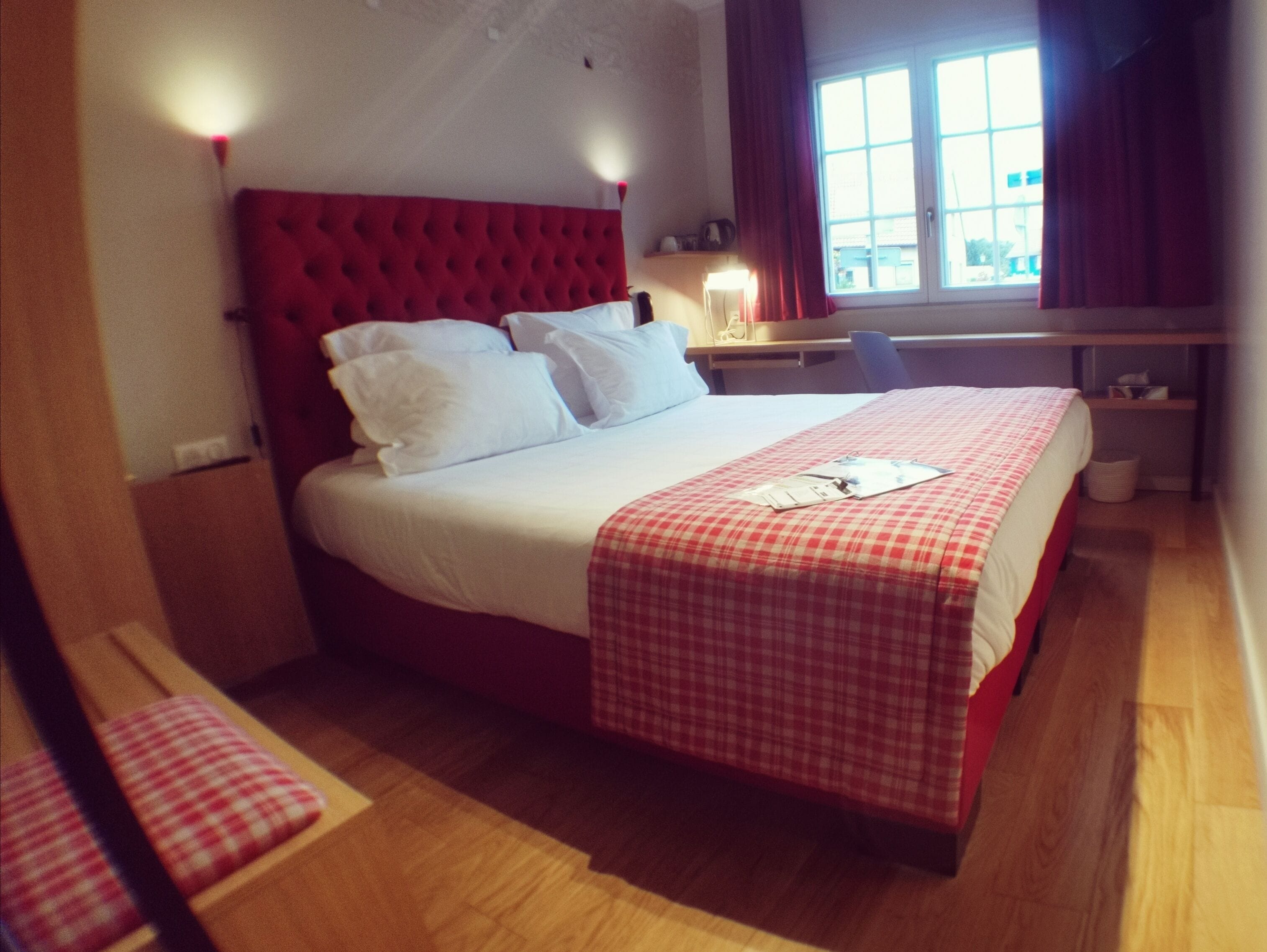 Ein Hotelzimmer mit rotem, gepolsterten Kopfteil, einem Bett mit weißer Bettwäsche, einer rot-weißen Karopfteppichdecke, einem Schreibtisch mit Lampe und einem Fenster mit Vorhängen.
