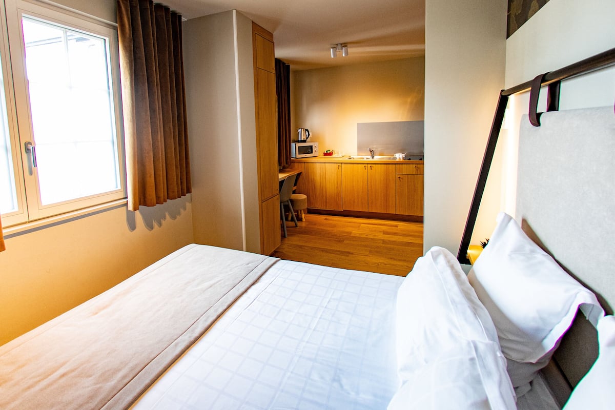 Habitación de hotel con cama, ventana con cortinas, kitchenette con microondas y suelo de madera.