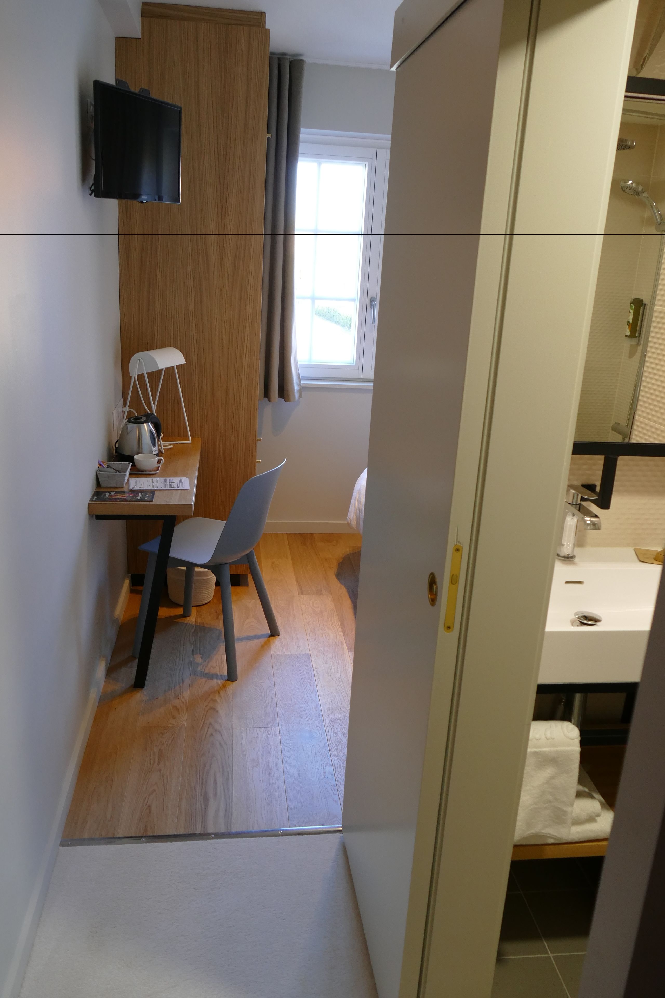 Quarto de hotel moderno com uma secretária de madeira, uma cadeira, uma televisão de tela plana na parede, uma janela com cortinas e um banheiro com pia e espelho.