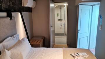 Camera d'albergo con un letto, una panca di legno, zona doccia e una porta con un numero.