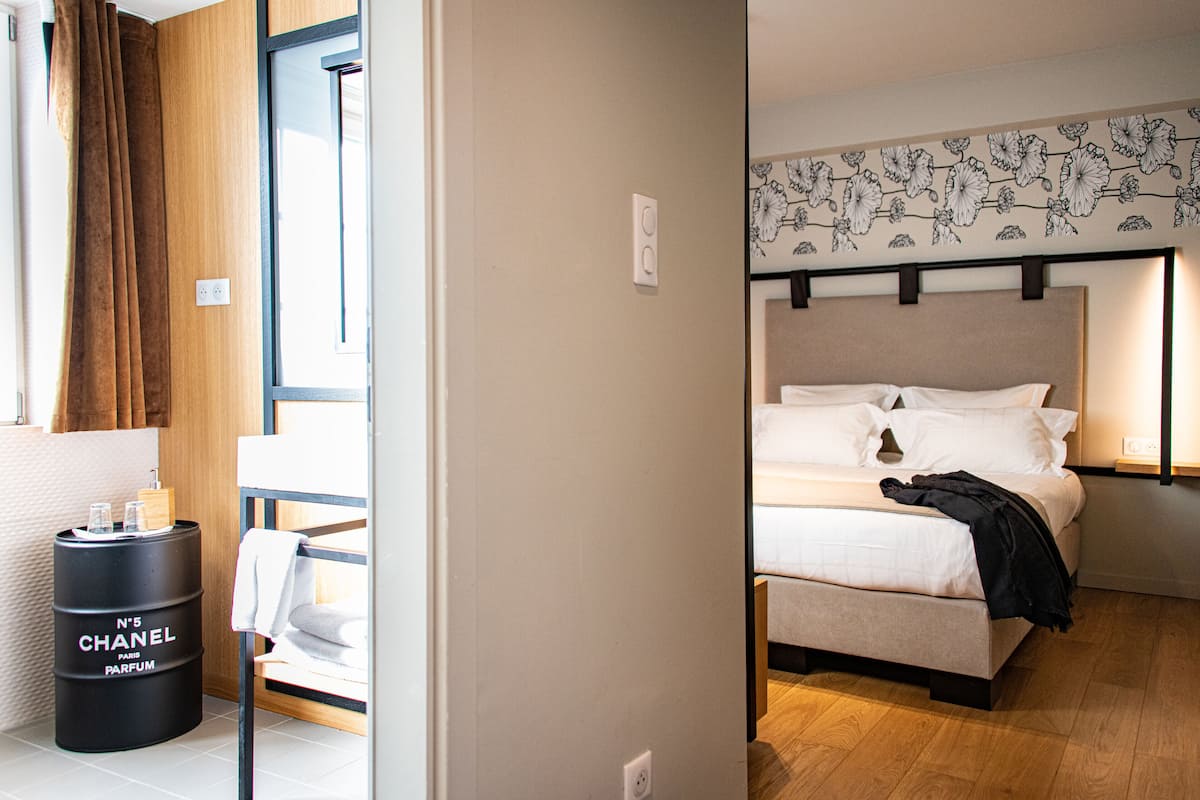 Una habitación de hotel con cama, mesita de noche, un frasco negro con el texto 'N°5 CHANEL PARIS PARFUM' y una ventana con cortinas.