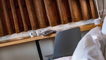 A hotel room with a wooden desk, a laptop, a white mug, a magazine, and a grey chair.