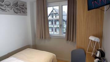 Ein Schlafzimmer mit Bett, einem Fenster mit Vorhängen, einem an der Wand befestigten Fernseher und einem Holztisch mit Wasserkocher und Tasse.