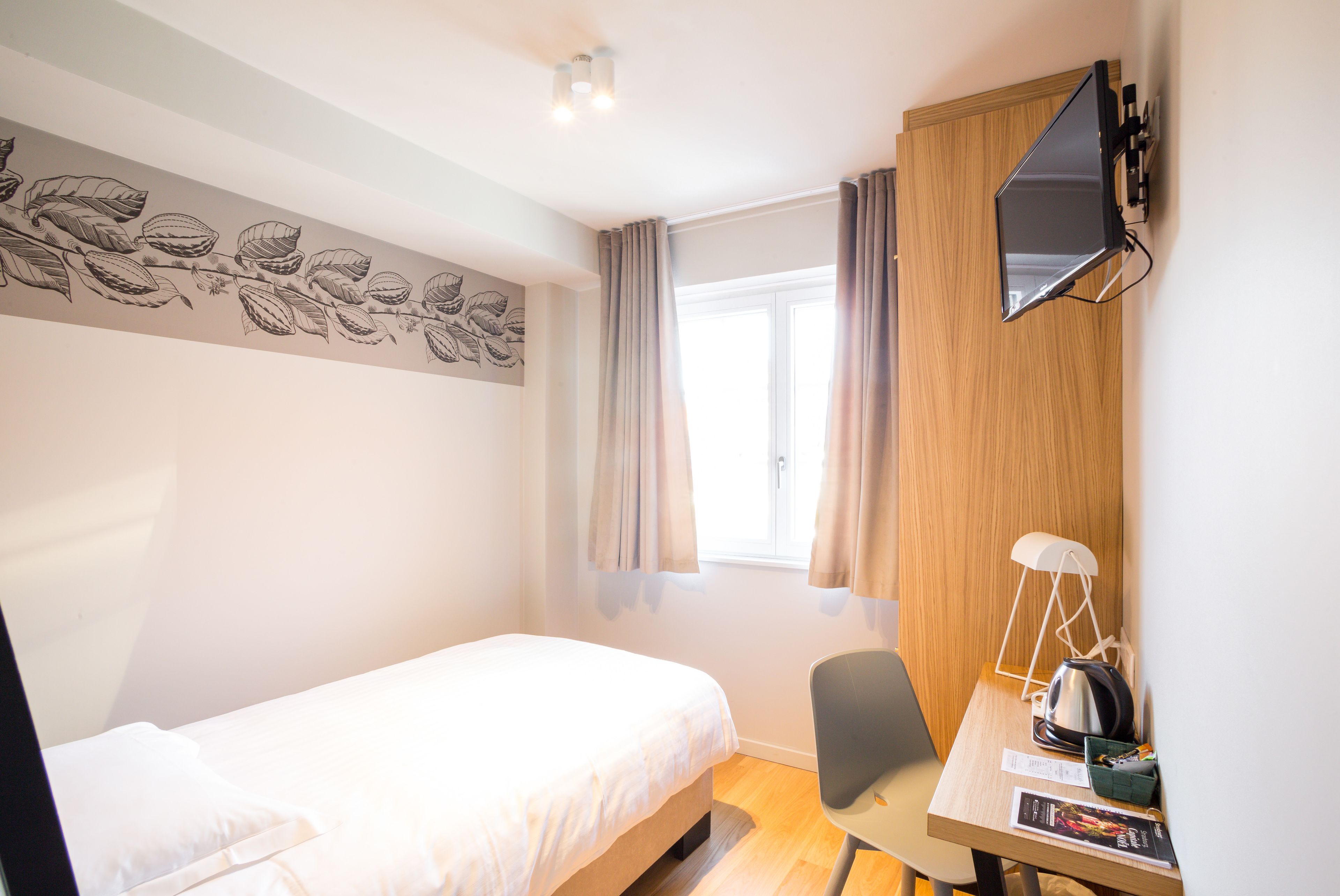 A bedroom with a bed, a desk, a chair, a flat-screen TV, and a window with curtains.