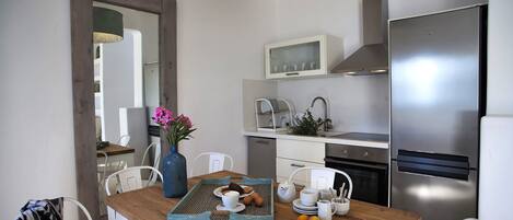 A modern kitchen with a wooden dining table, white chairs, a blue vase with flowers, and a stainless steel refrigerator.