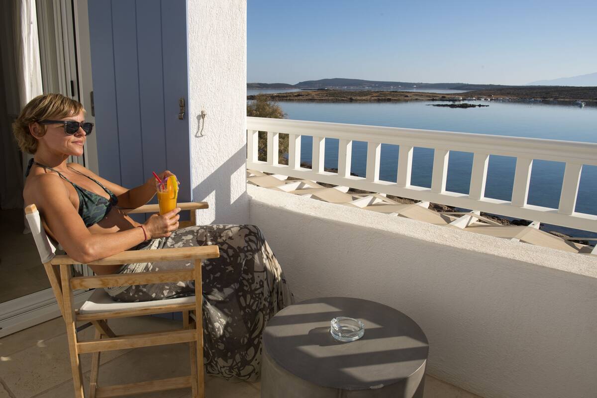 A person seated on a wooden chair on a balcony with a view of a calm lake and distant hills.