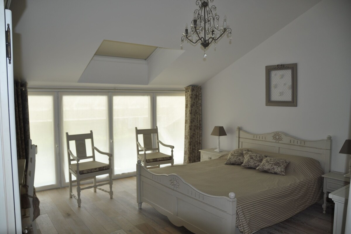 Ein Schlafzimmer mit einem großen Bett, zwei Sessel, einem Nachttisch, einer Lampe, einem Kronleuchter, einem Fenster mit Vorhängen und einem gerahmten Bild an der Wand.