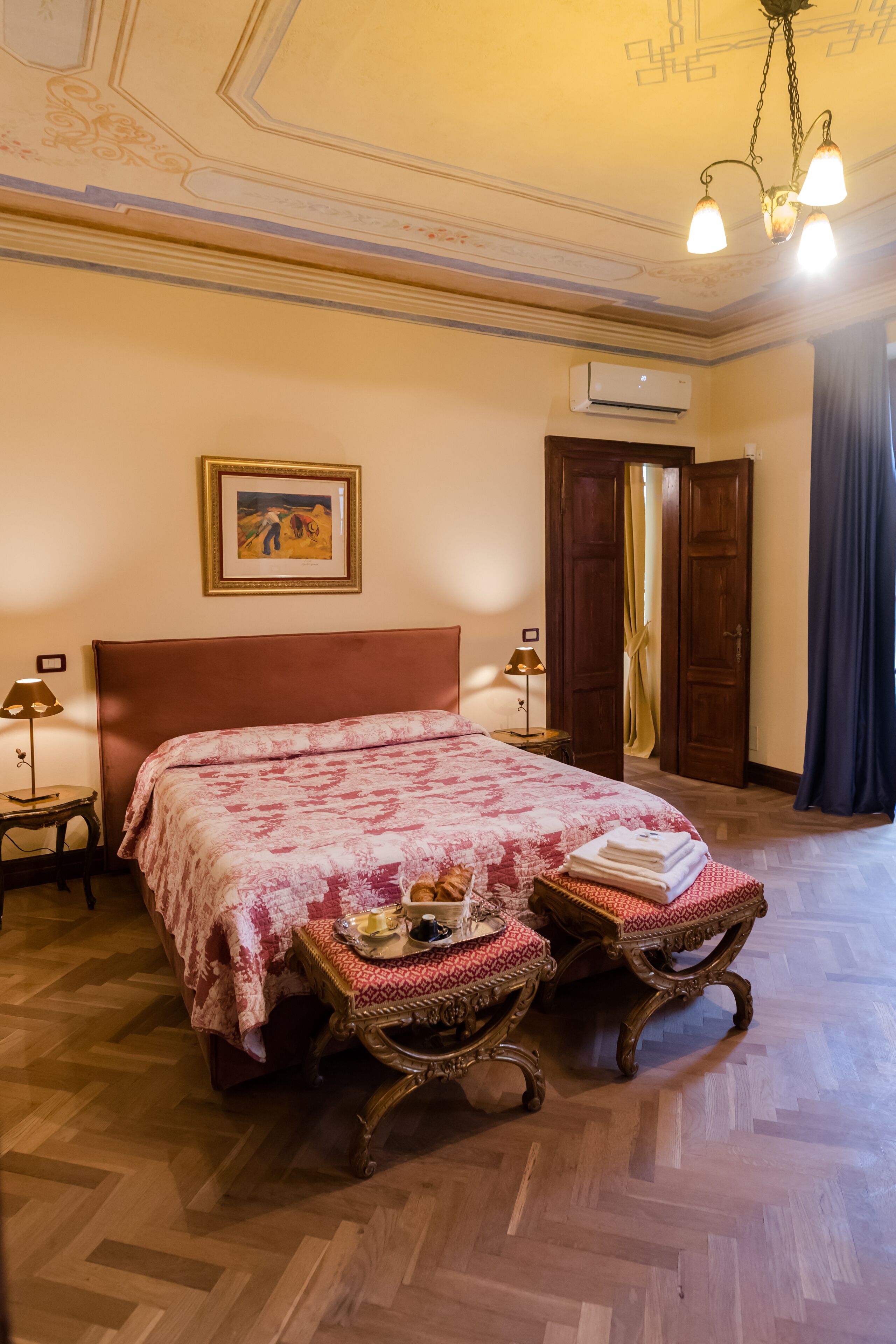 Ein Hotelzimmer mit einem großen Bett, Holzboden, einer Kristallleuchtre, einer Klimaanlage und einer Tür mit Vorhängen.