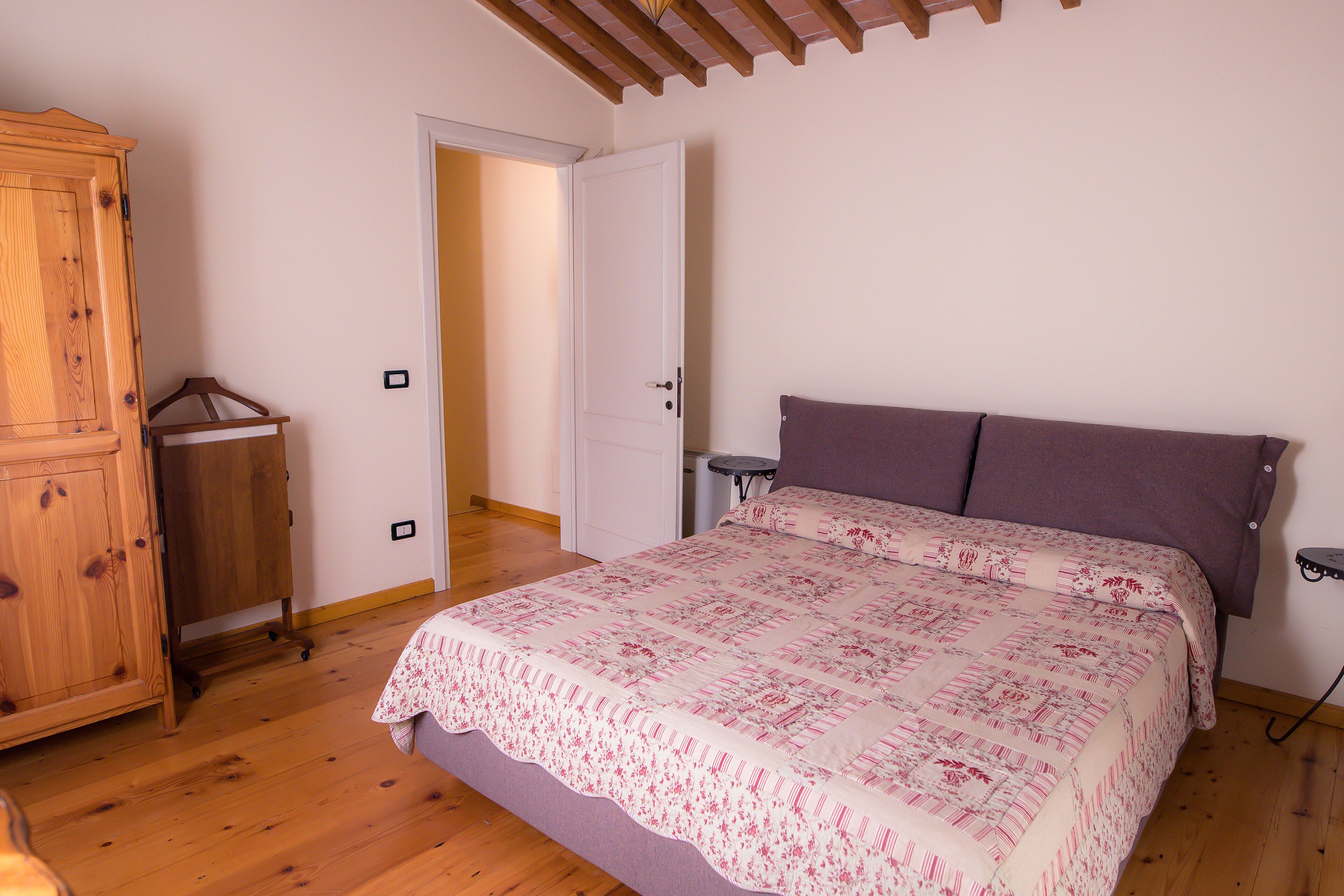 Un dormitorio con un armario de madera, una cama con colcha estampada y una puerta que da a otra habitación.