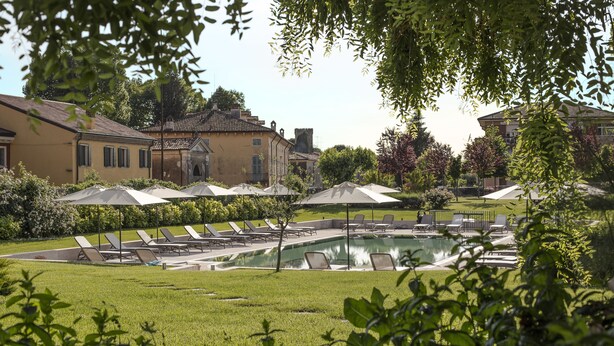 A pool area with lounge chairs and umbrellas, surrounded by greenery and buildings. - Villa Nichesola (Colognola ai Colli)