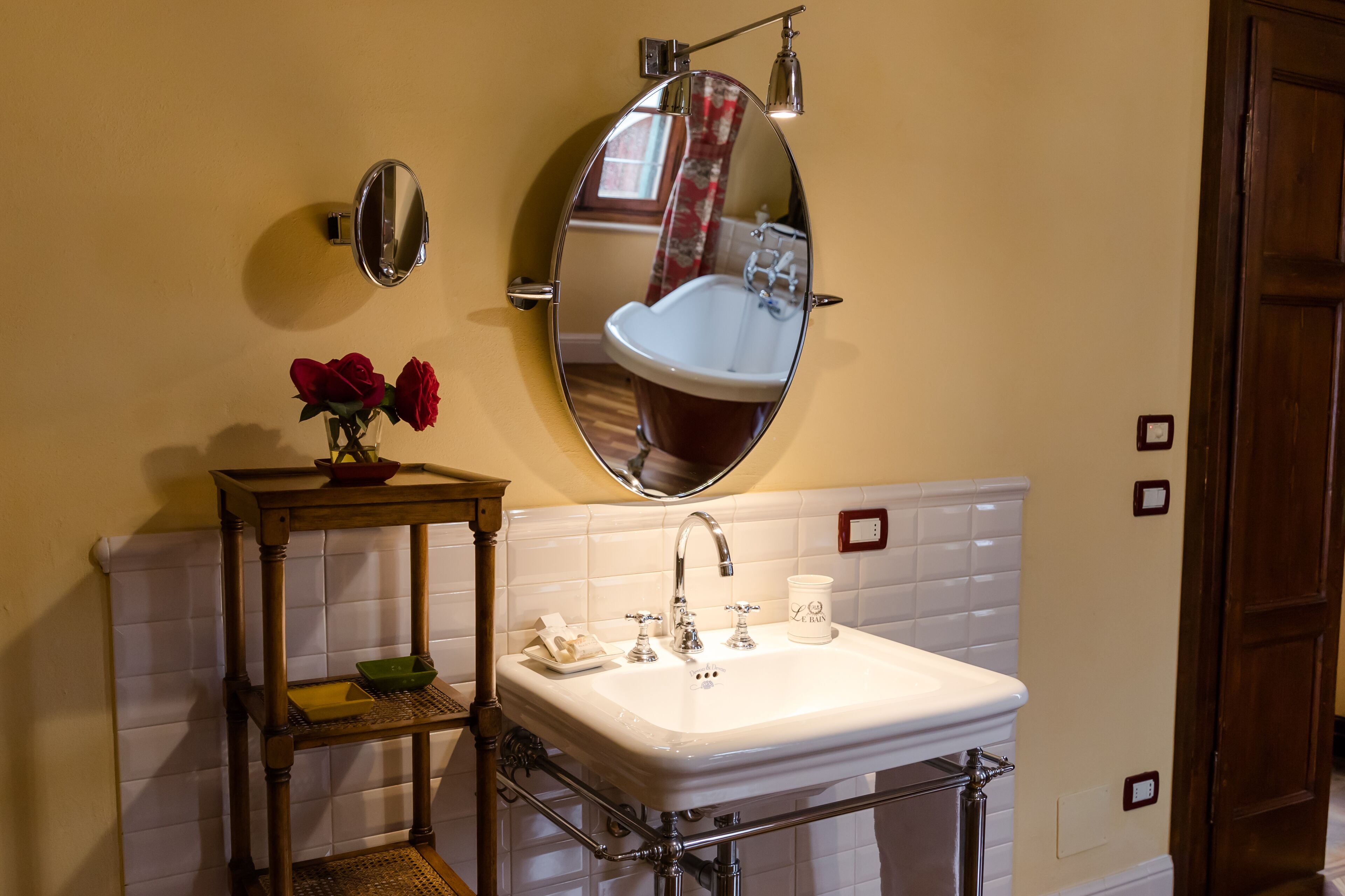A bathroom with a white sink, a mirror, and a wooden shelf with a vase of red roses.