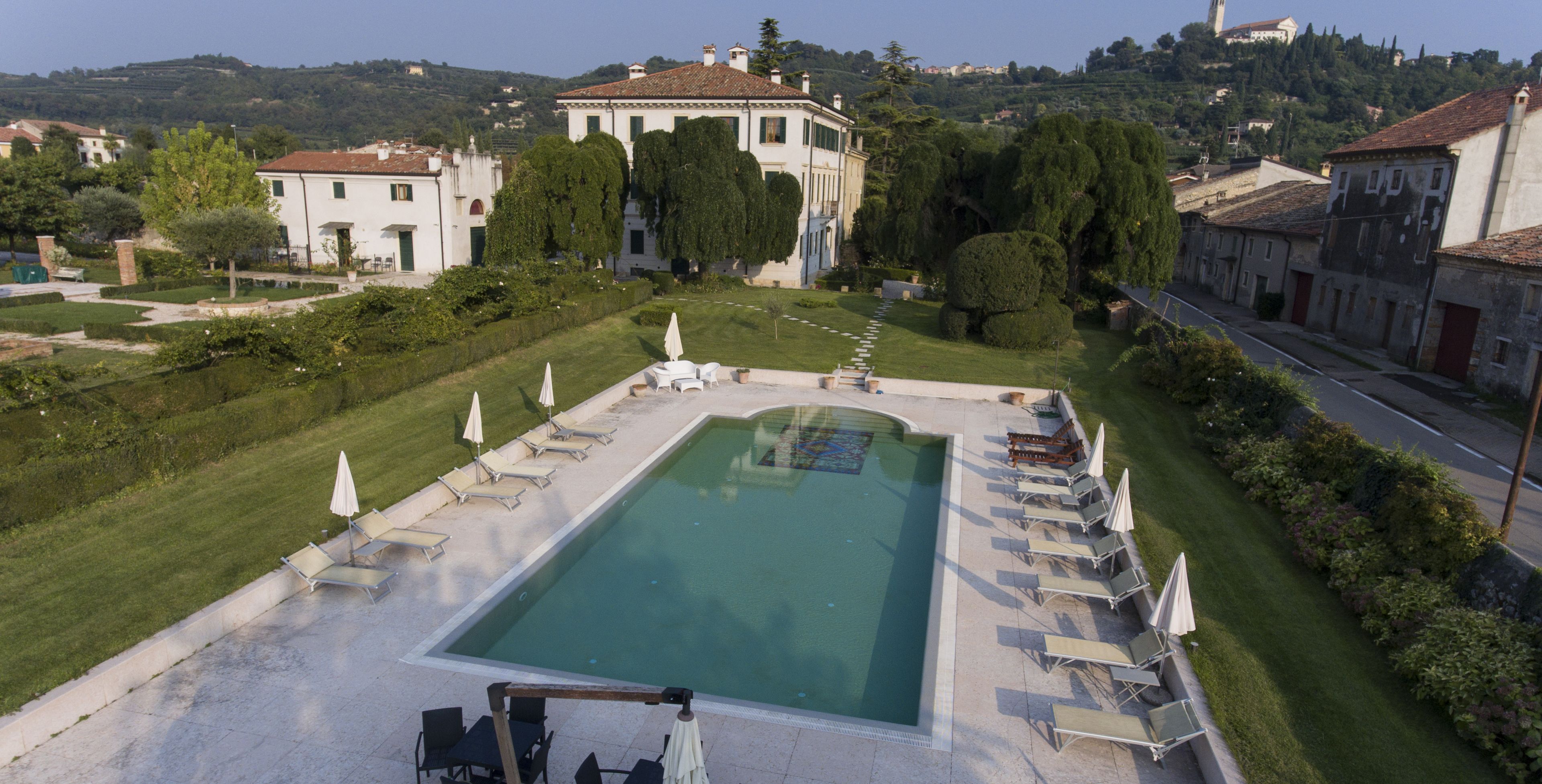 A rectangular swimming pool with lounge chairs and umbrellas, surrounded by well-maintained lawns and trees, with buildings in the background.