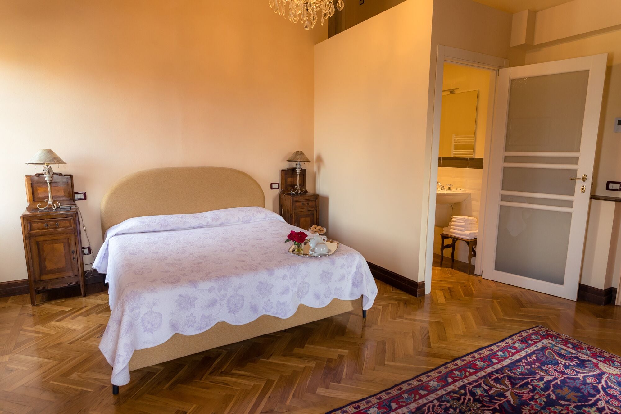 Ein Schlafzimmer mit einem Bett, zwei Nachttischen mit Lampen, einem kleinen Tisch mit Blumenvase und einem durchgehenden Türrahmen sichtbaren Badezimmer.