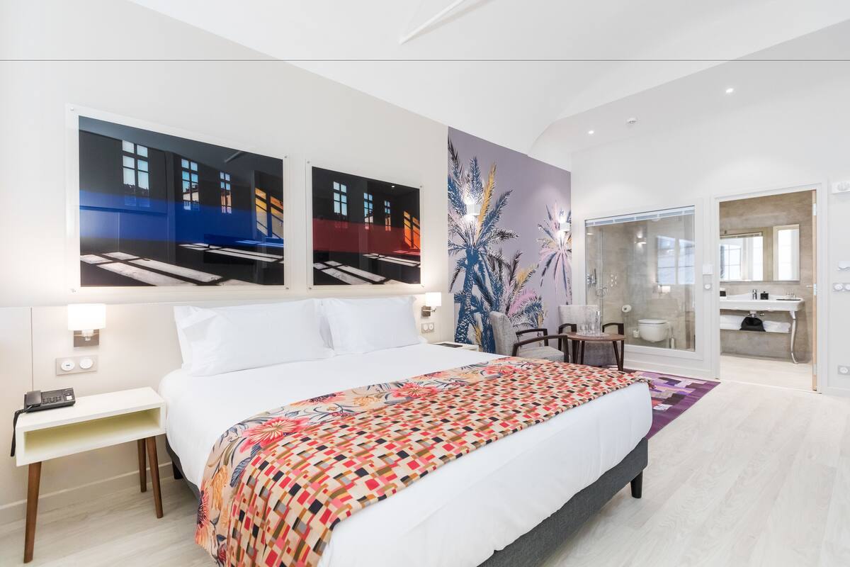 Une chambre d’hôtel moderne, dotée d’un grand lit, d’un bureau, d’une chaise et d’une salle de bain visible à travers une porte ouverte.