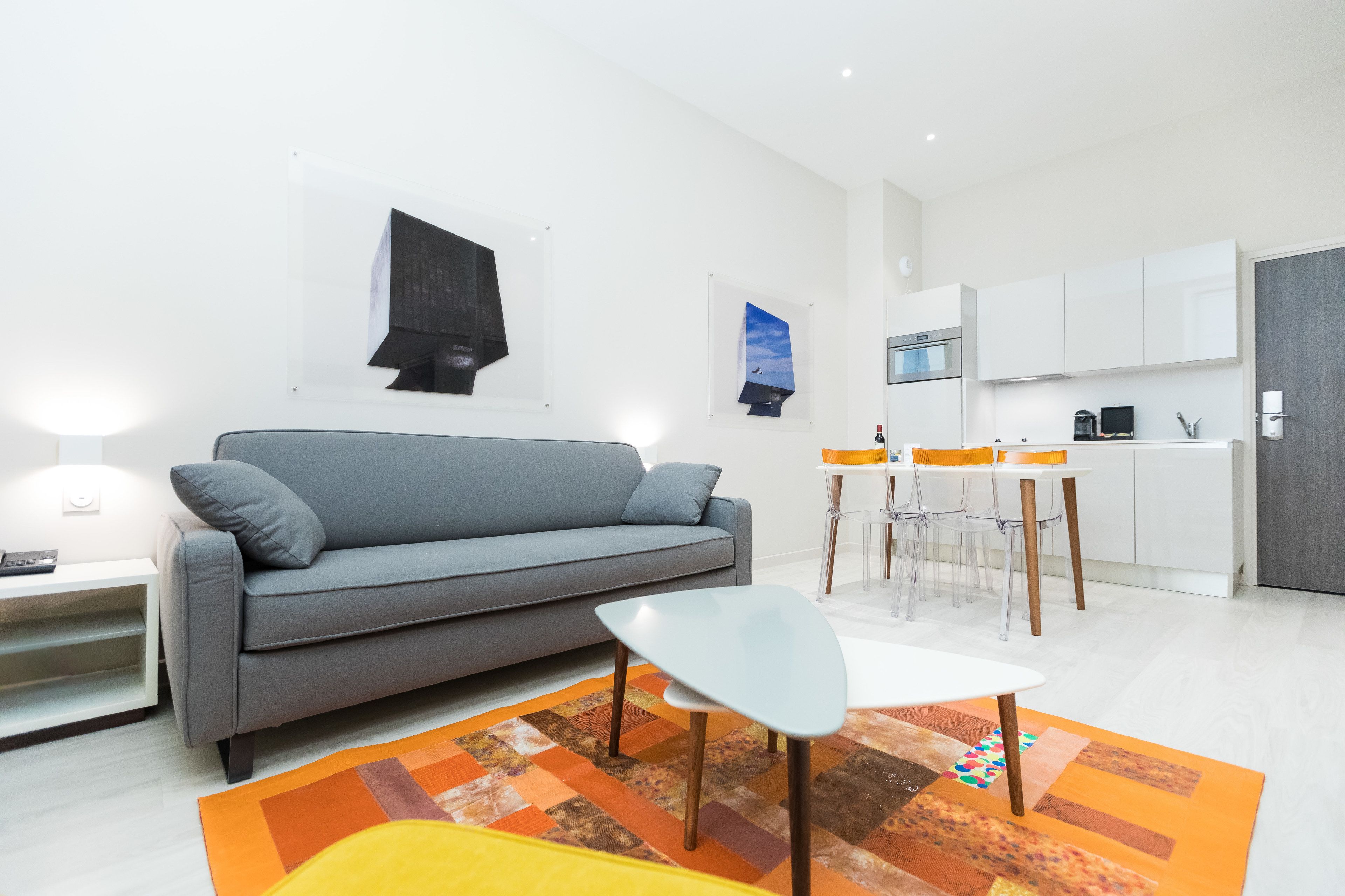 Una sala de estar moderna con un sofá gris, una mesa de centro blanca y un tapete colorido. Hay una zona de cocina con armarios blancos y una mesa de comedor con sillas.