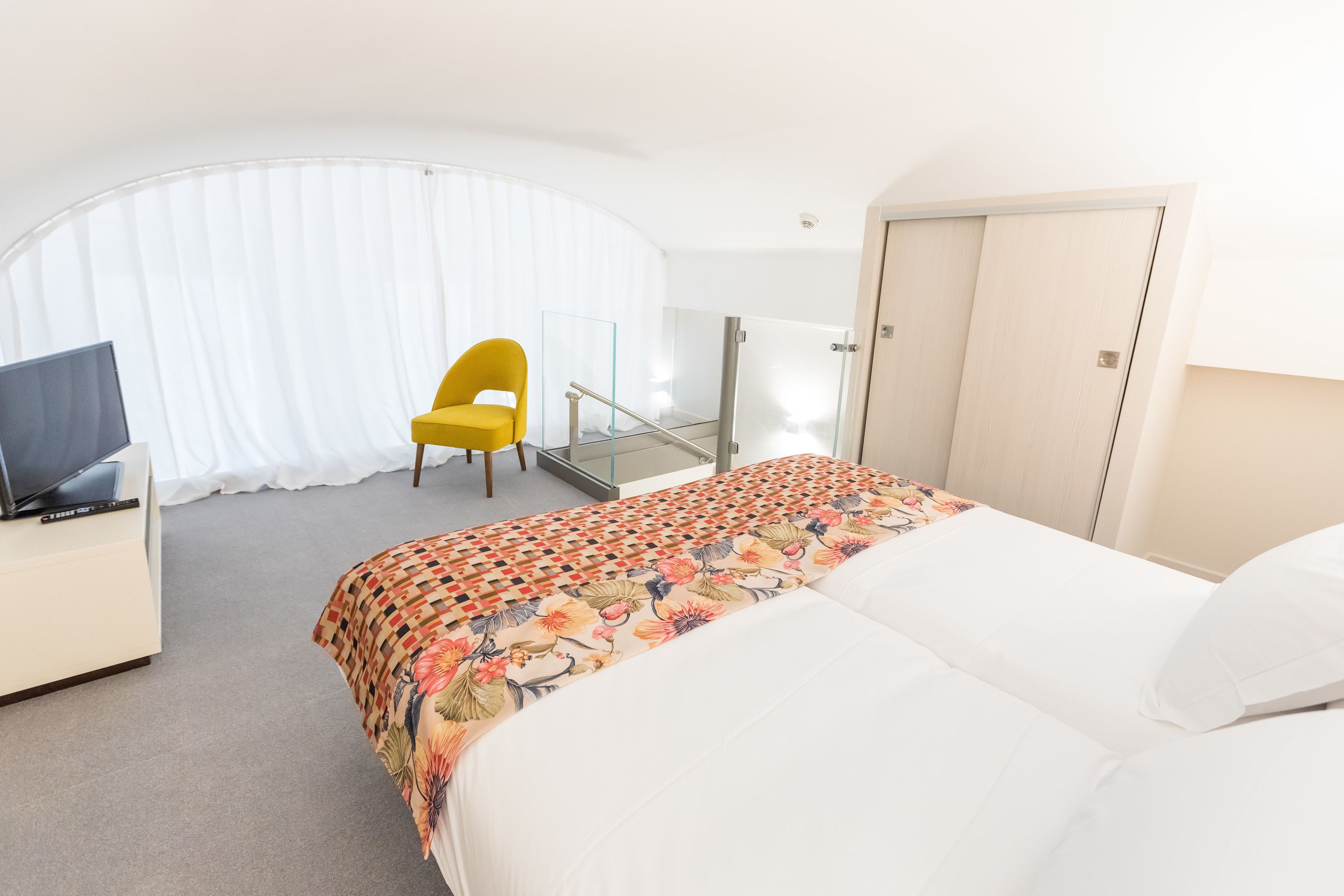 A hotel room with a bed, a chair, a TV, and a glass door leading to a balcony.
