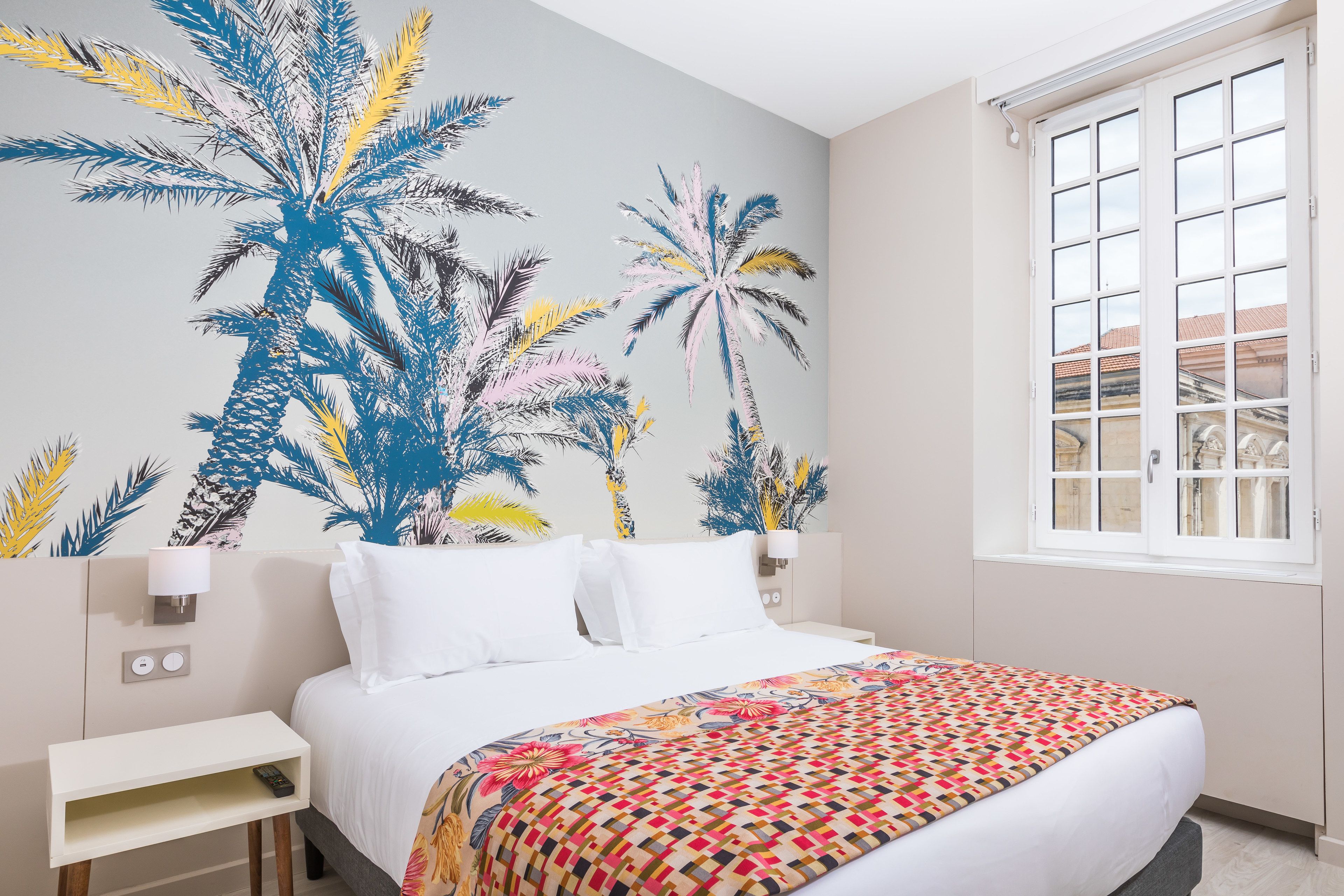 A bedroom with a bed, white bedding, a colorful geometric-patterned blanket, a wall mural of palm trees, a bedside table with a lamp, and a window with a view of buildings.
