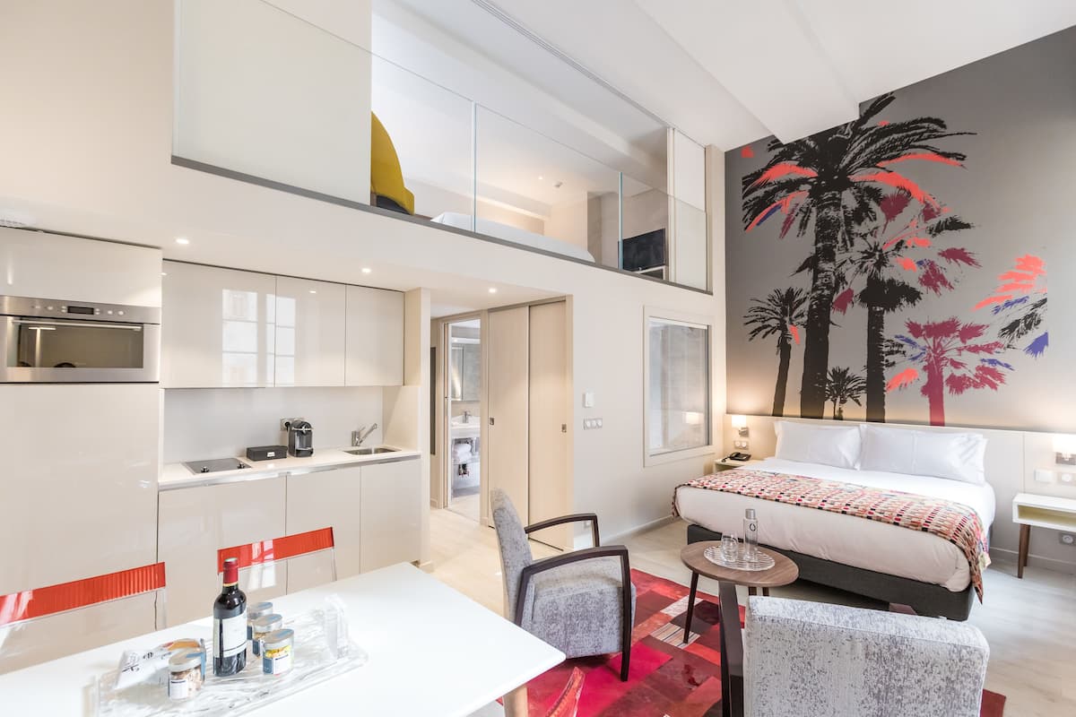 Une chambre d’hôtel moderne comprenant un lit, un coin repas avec une table et des chaises, un coin cuisine avec un four à micro-ondes et une machine à café, ainsi qu’une grande fresque murale.
