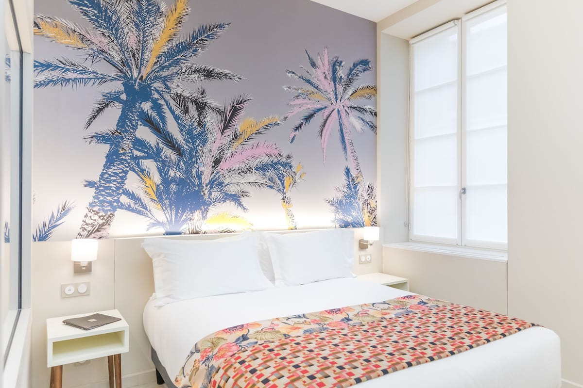 Une chambre d’hôtel avec un lit, une table de chevet, un mur orné d’une fresque représentant un palmier et une fenêtre.
