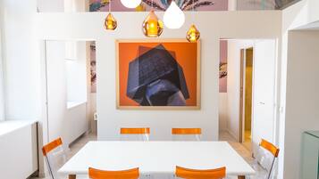 Un comedor moderno con una mesa blanca, seis sillas transparentes con detalles naranja y luces colgantes.