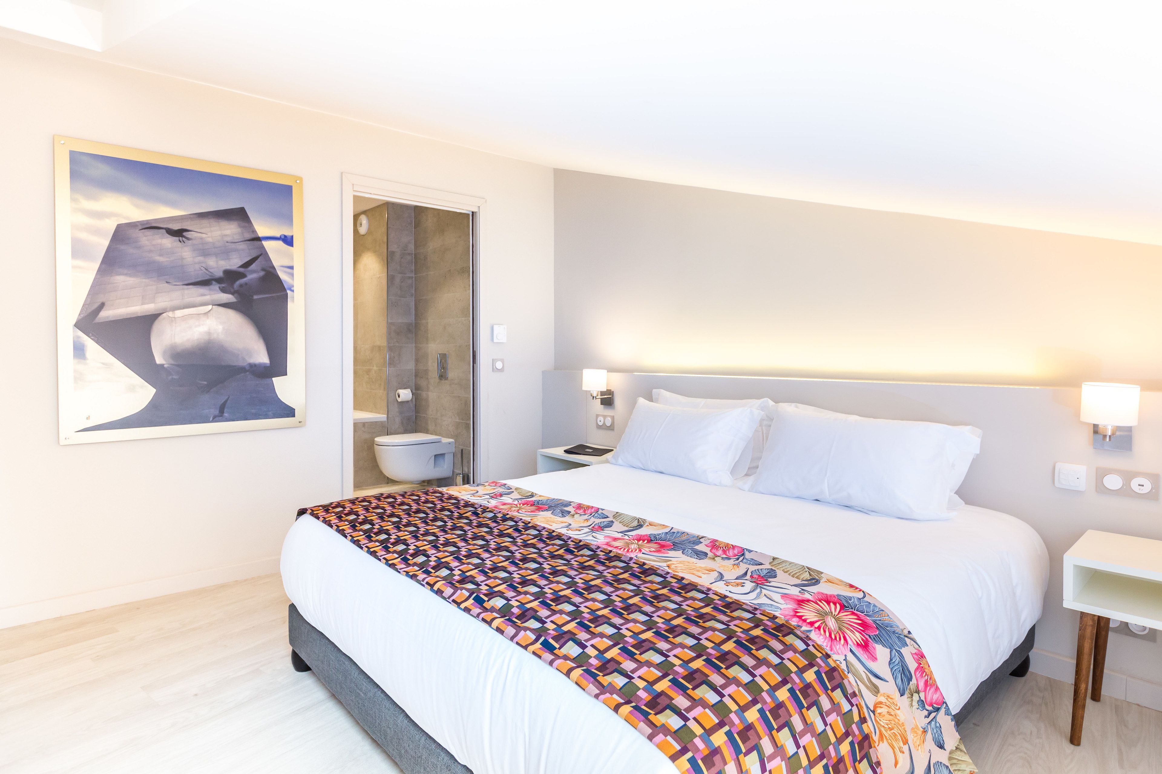A modern hotel room with a large bed, a bathroom with a glass shower, and a wall-mounted artwork.
