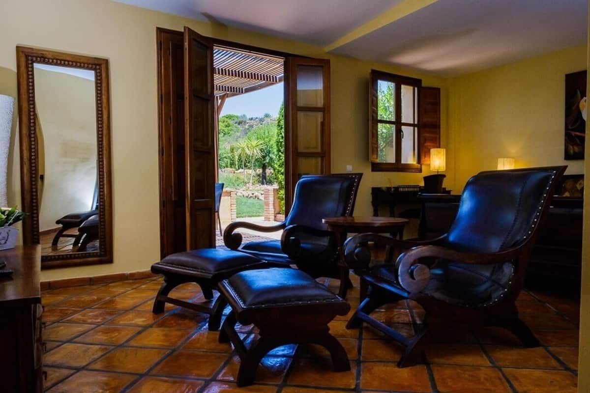 Una habitación con piso de baldosas, dos sillones de cuero, una mesa de madera, un espejo y una ventana con vista a vegetación.