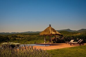 Un quiosco con techo de paja, con piscina y sillones de descanso, en un paisaje verde y exuberante.