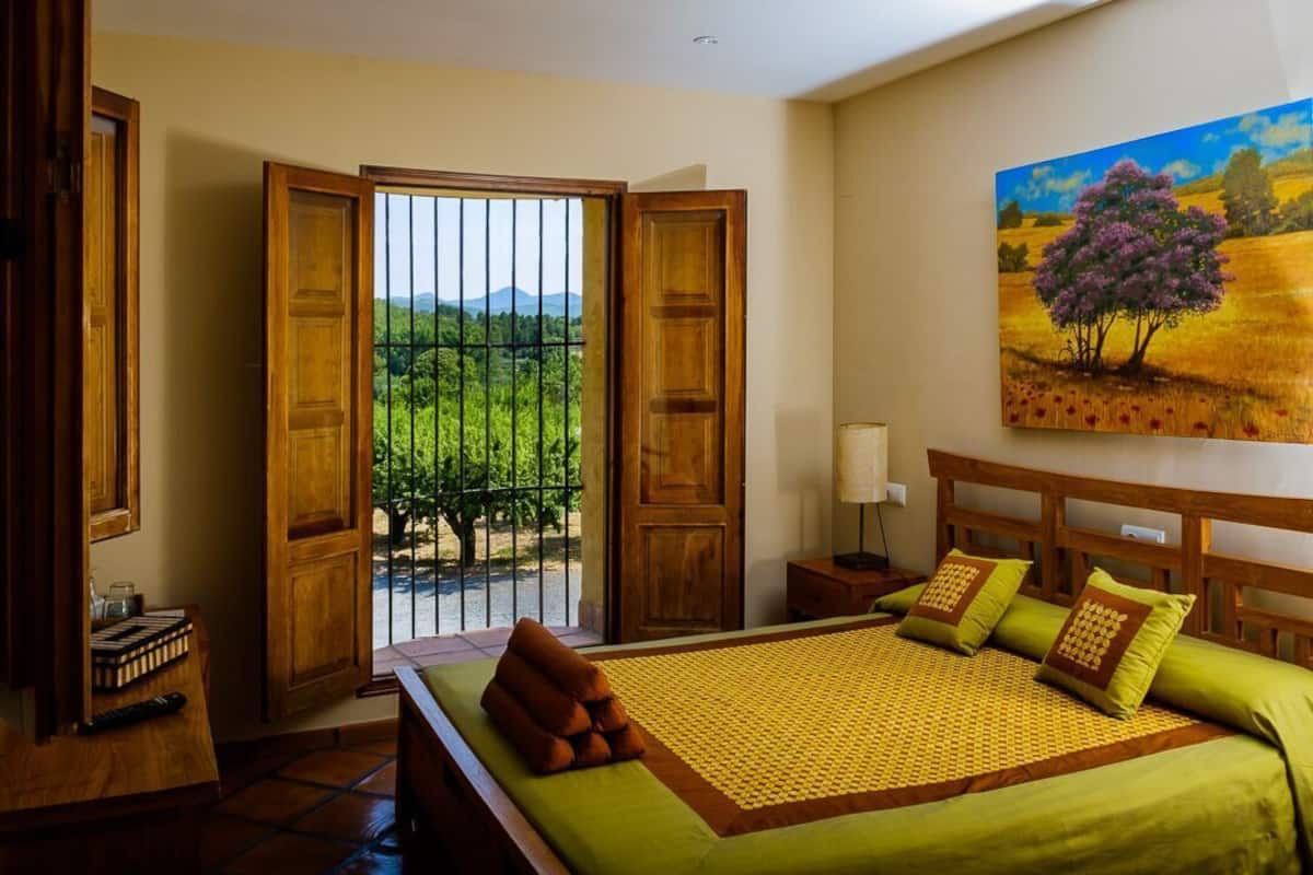 Un dormitorio con cama, ropa de cama verde, cabecera de madera, mesita de noche, lámpara y un cuadro de un árbol.