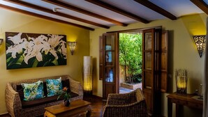 Una sala de estar acogedora con un sofá de mimbre, una mesa de centro de madera y un cuadro de lirios blancos. Hay dos apliques de pared y un jarrón alto con ramas.