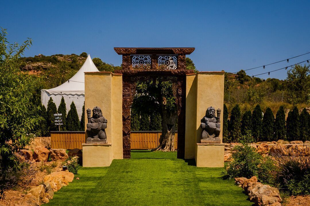 A garden entrance with a decorative archway, a white tent, and statues on either side.