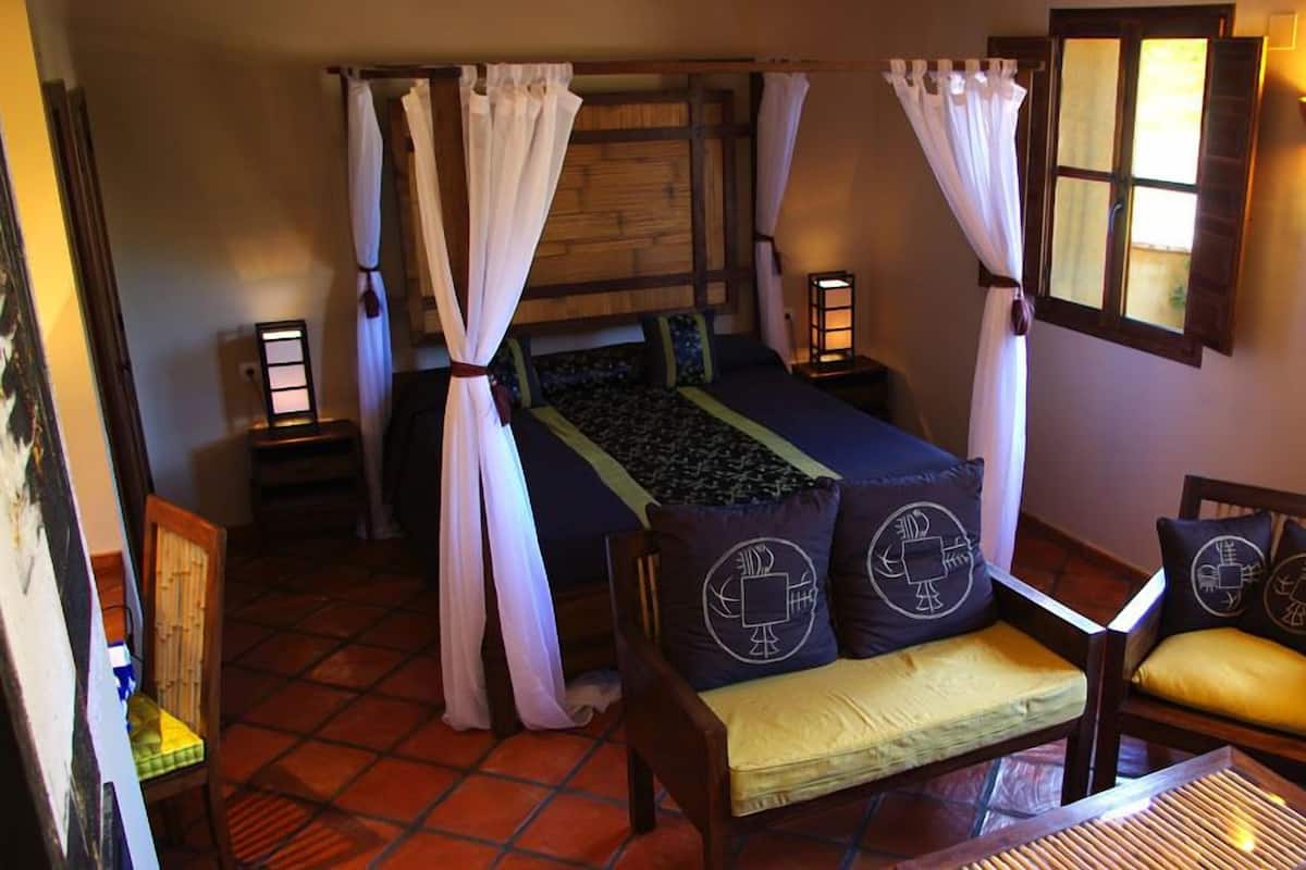 Una habitación con una cama, dos mesitas de noche, una silla y un ventanal con cortinas.