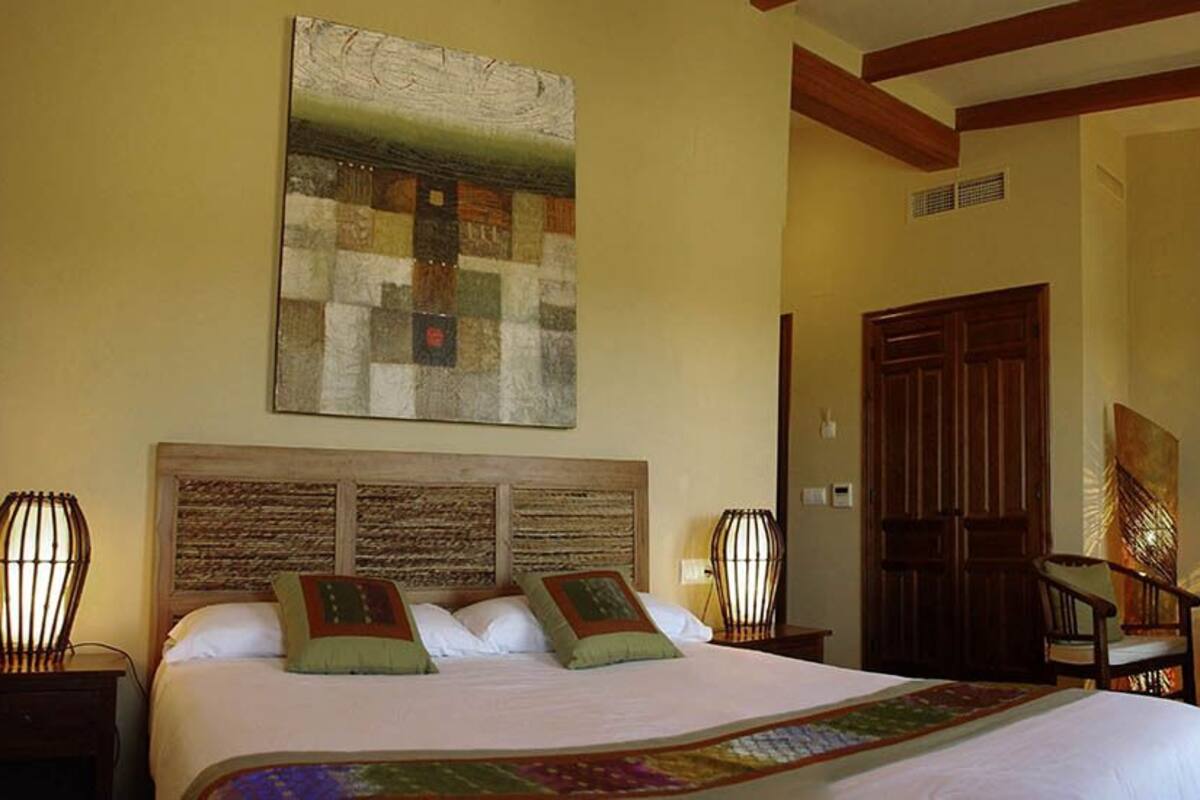 Habitación de hotel con una cama grande, dos lámparas de noche, un cabecero de madera, un cuadro en la pared, una puerta de madera y una pequeña mesa con sillas.