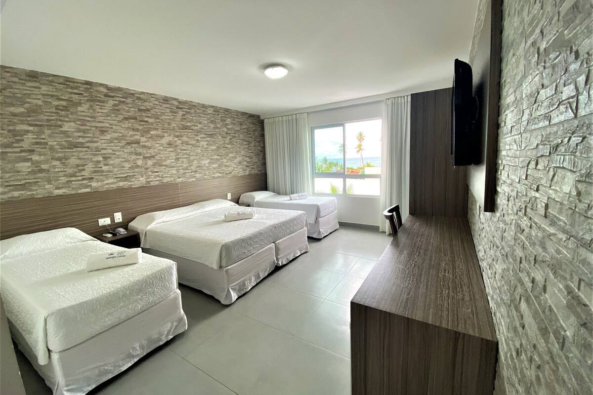 Een hotelkamer met drie bedden, een aan de muur bevestigde televisie, een raam met uitzicht op het strand en een muur met een stenen textuur.