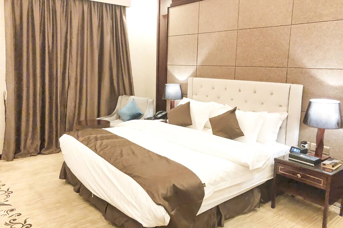 A hotel room with a large bed, a chair, a nightstand with a lamp, and a curtain.