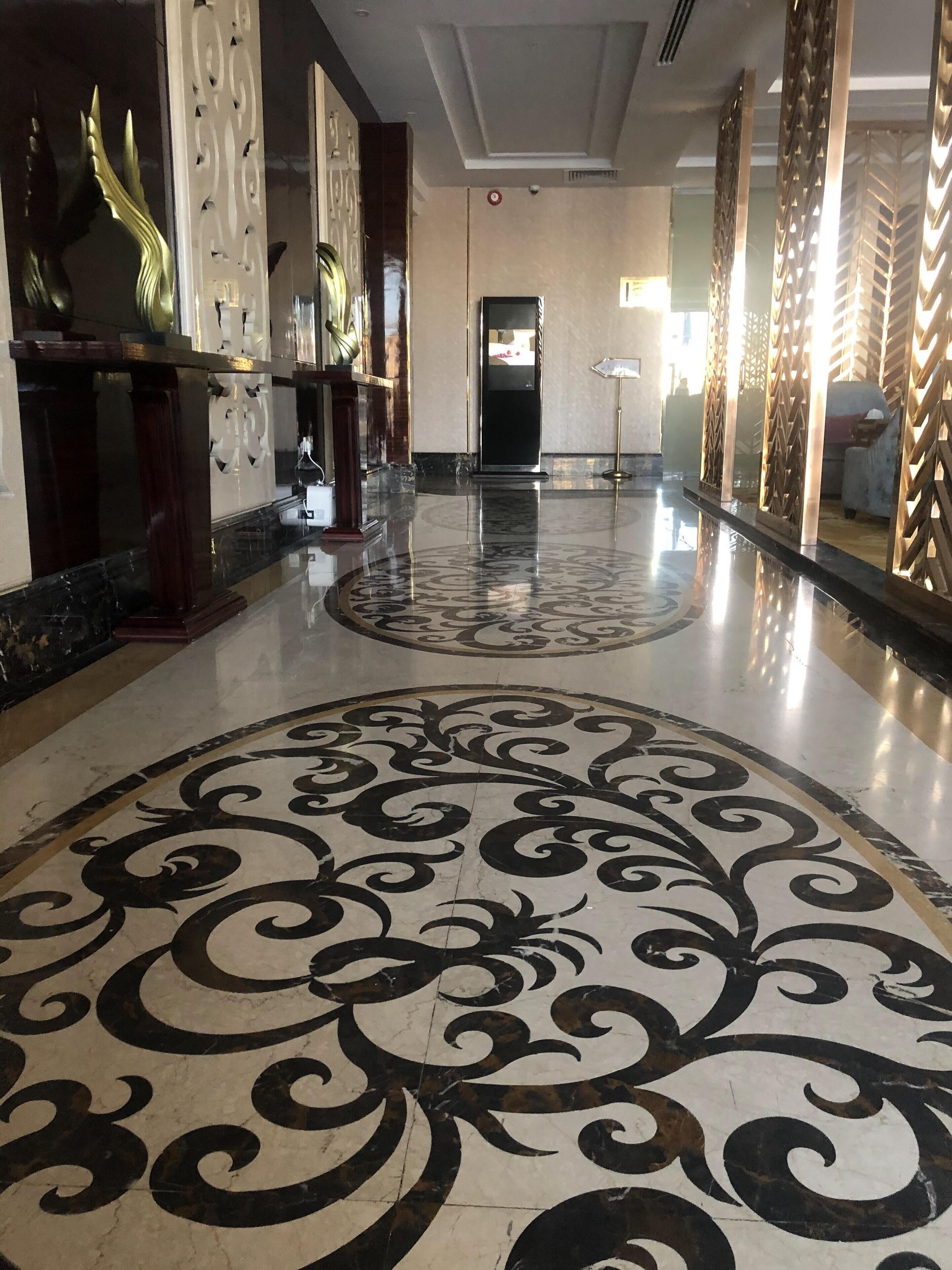A lobby with a polished marble floor featuring a black and white pattern, a reception desk with a decorative backdrop, and a modern interior design with a high ceiling and patterned wall panels.