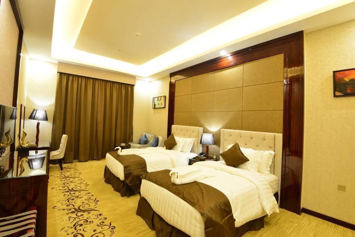 A hotel room with two beds, a desk with a lamp, a chair, and a large window with curtains.