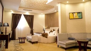 A hotel room with a curved bed, a sofa, a small table, and a painting on the wall. - Seven Garden AL wadi (Riyadh)