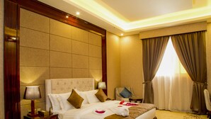 A hotel room with a large bed, a nightstand with a lamp, a chair, and a window with curtains. - Seven Garden AL wadi (Riyadh)