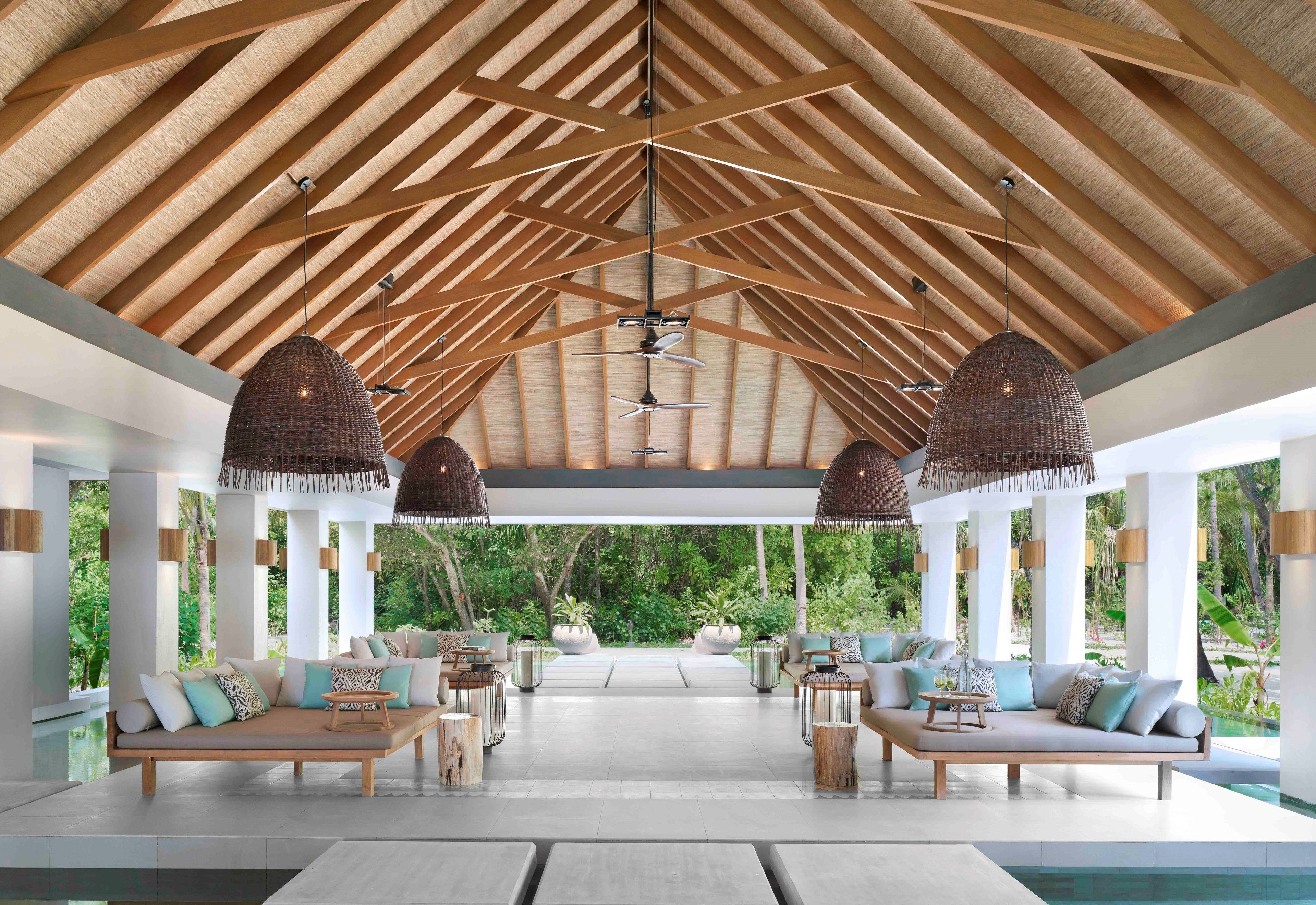 A spacious indoor area with a high wooden ceiling, large hanging wicker lamps, and a pool in the background.