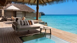 Des bungalows sur pilotis dotés d’une terrasse en bois, de chaises longues et d’une piscine à débordement avec un fond en verre.