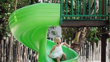 A green spiral slide at a playground with a wooden fence and trees in the background.
