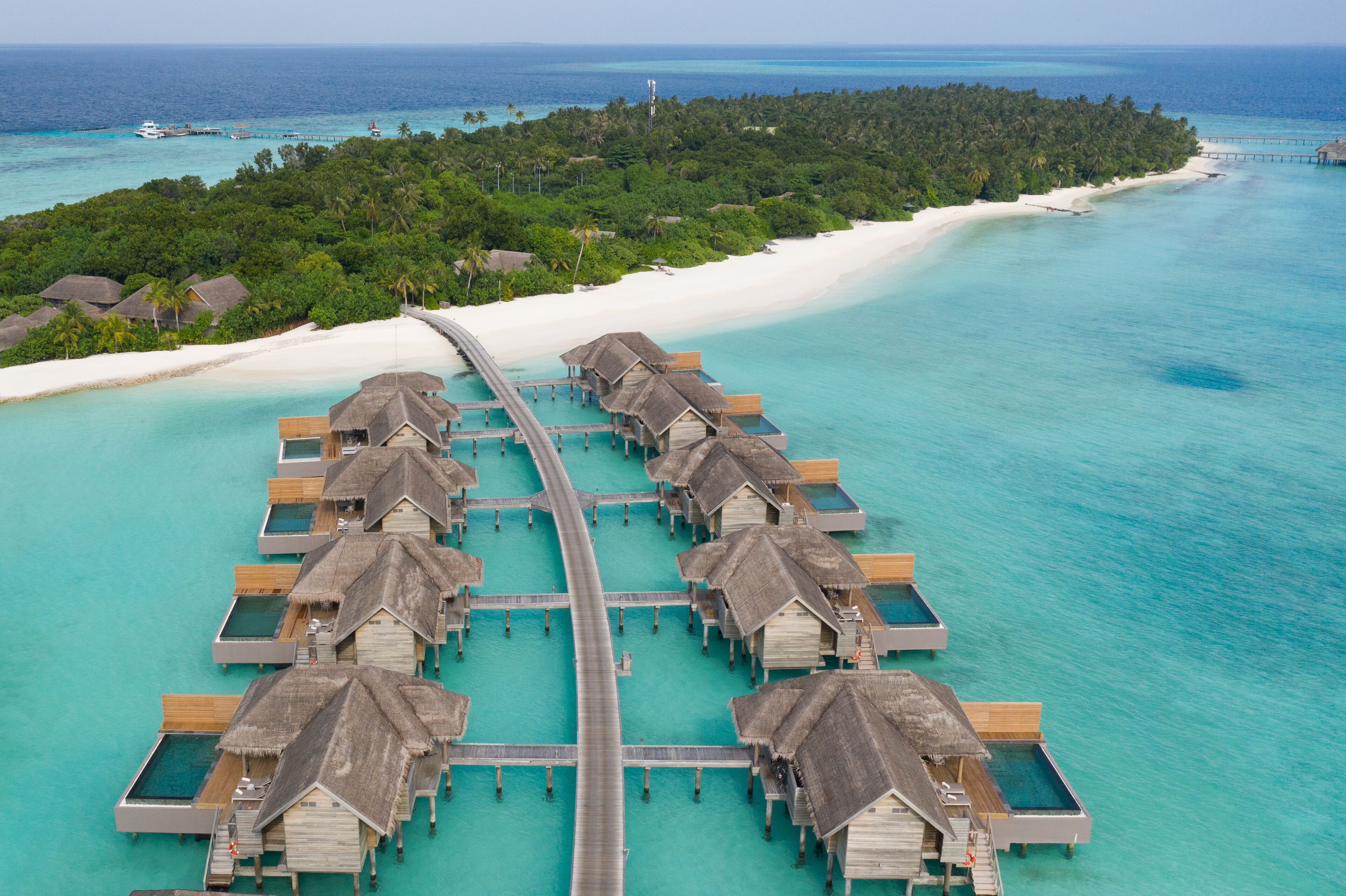 An aerial view of overwater bungalows connected by a walkway over turquoise waters, with a white sandy beach and lush greenery in the background.