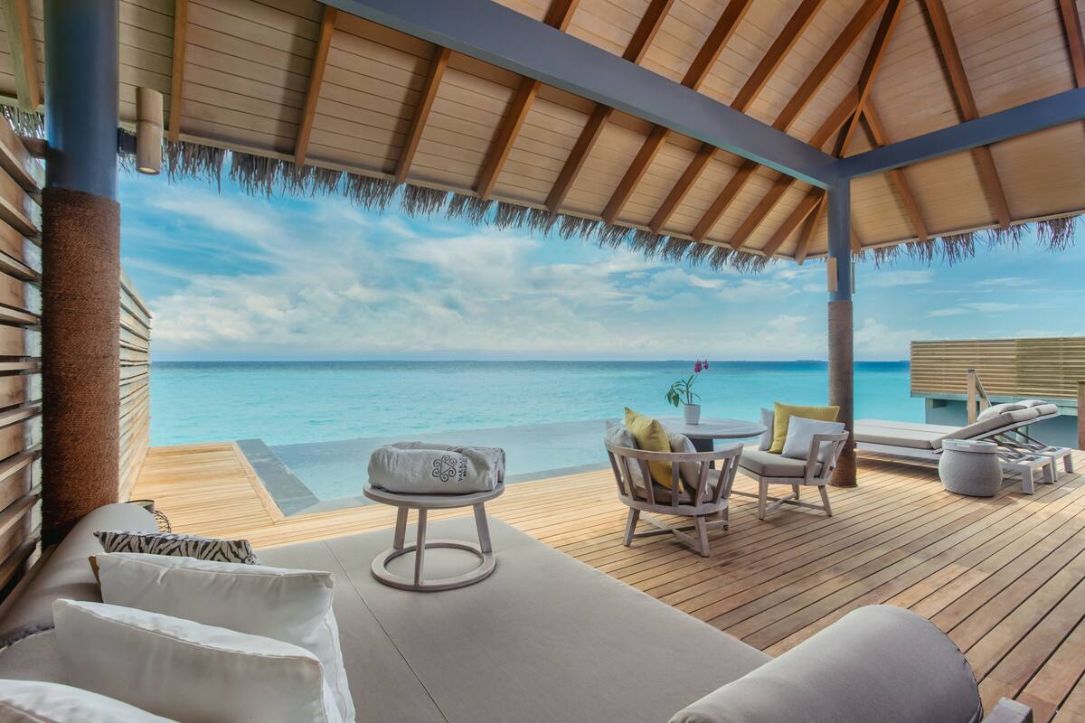 Terraza de madera con techo de paja, con vistas a un mar azul y transparente. Hay sillas de descanso y una mesa pequeña con cojines.