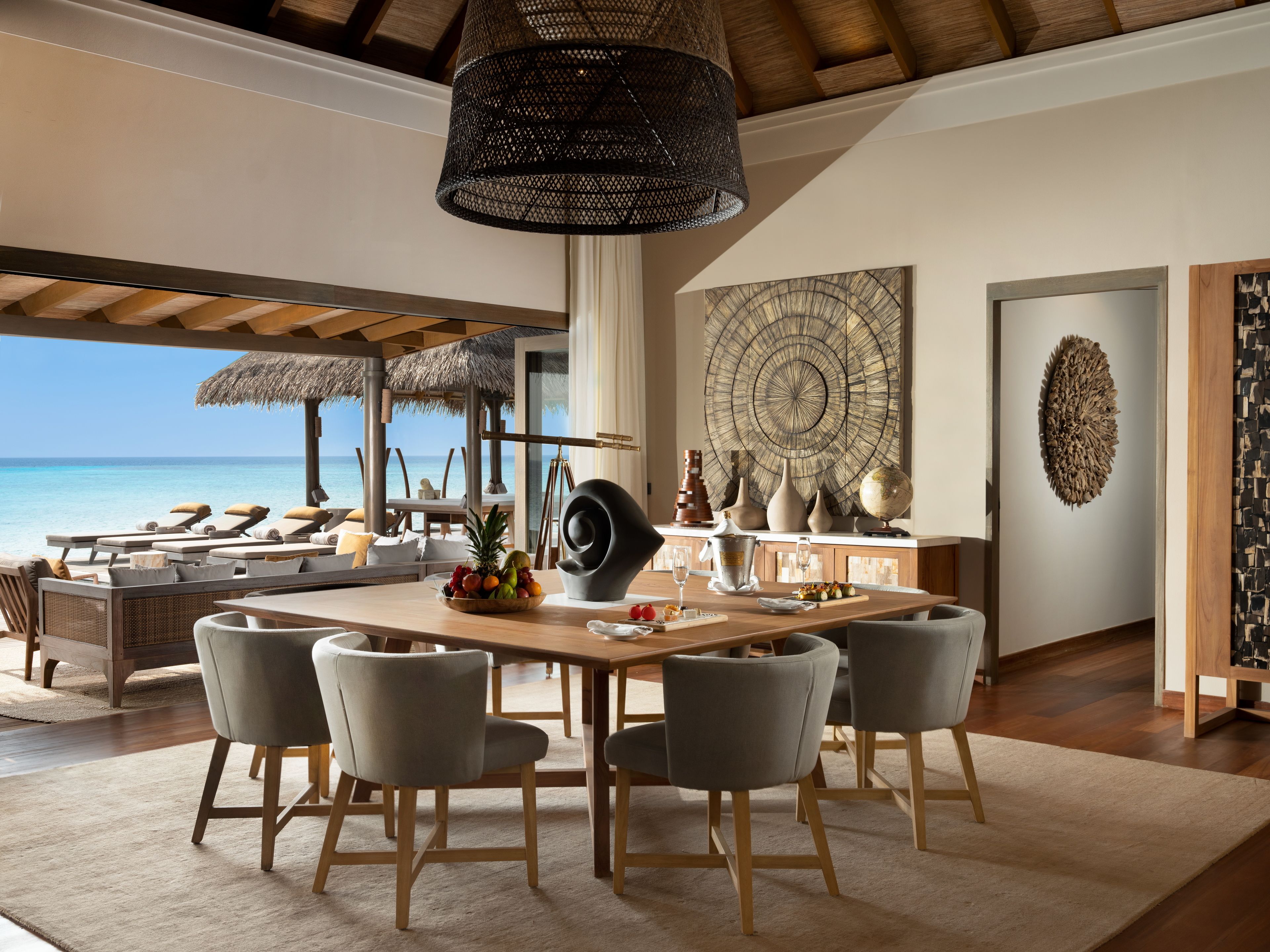 A beachside room with a wooden dining table, grey chairs, a fruit bowl, and a large wall art piece.