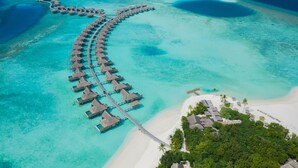Vue aérienne de bungalows sur pilotis reliés par une passerelle, d’une plage et d’un complexe hôtelier avec des bâtiments et des palmiers.
