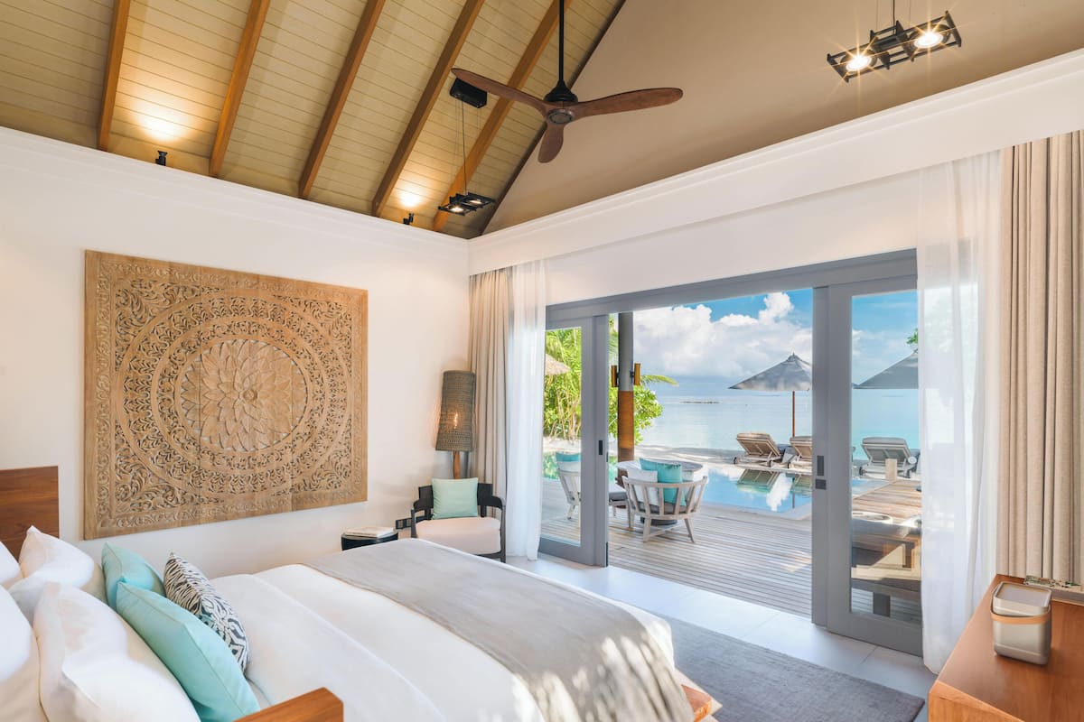 Un dormitorio con una cama grande, ventilador de techo y vistas a una piscina y una playa.
