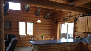 A cabin interior with a wooden dining area, a kitchen with wooden cabinets, and deer antlers mounted on the ceiling.