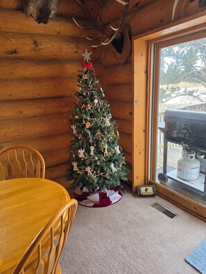 A wooden cabin interior with a Christmas tree, a wooden dining table, and a window with a view of the outdoors. - Family Friendly 4BR, 3 Bath Cabin w/Hot Tub in the heart of the Black Hills (Hill City)