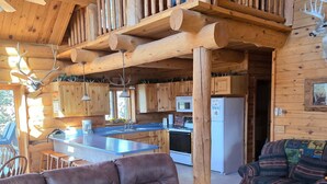 A log cabin interior with a wooden balcony, a kitchen area, and a brown leather sofa with decorative pillows.